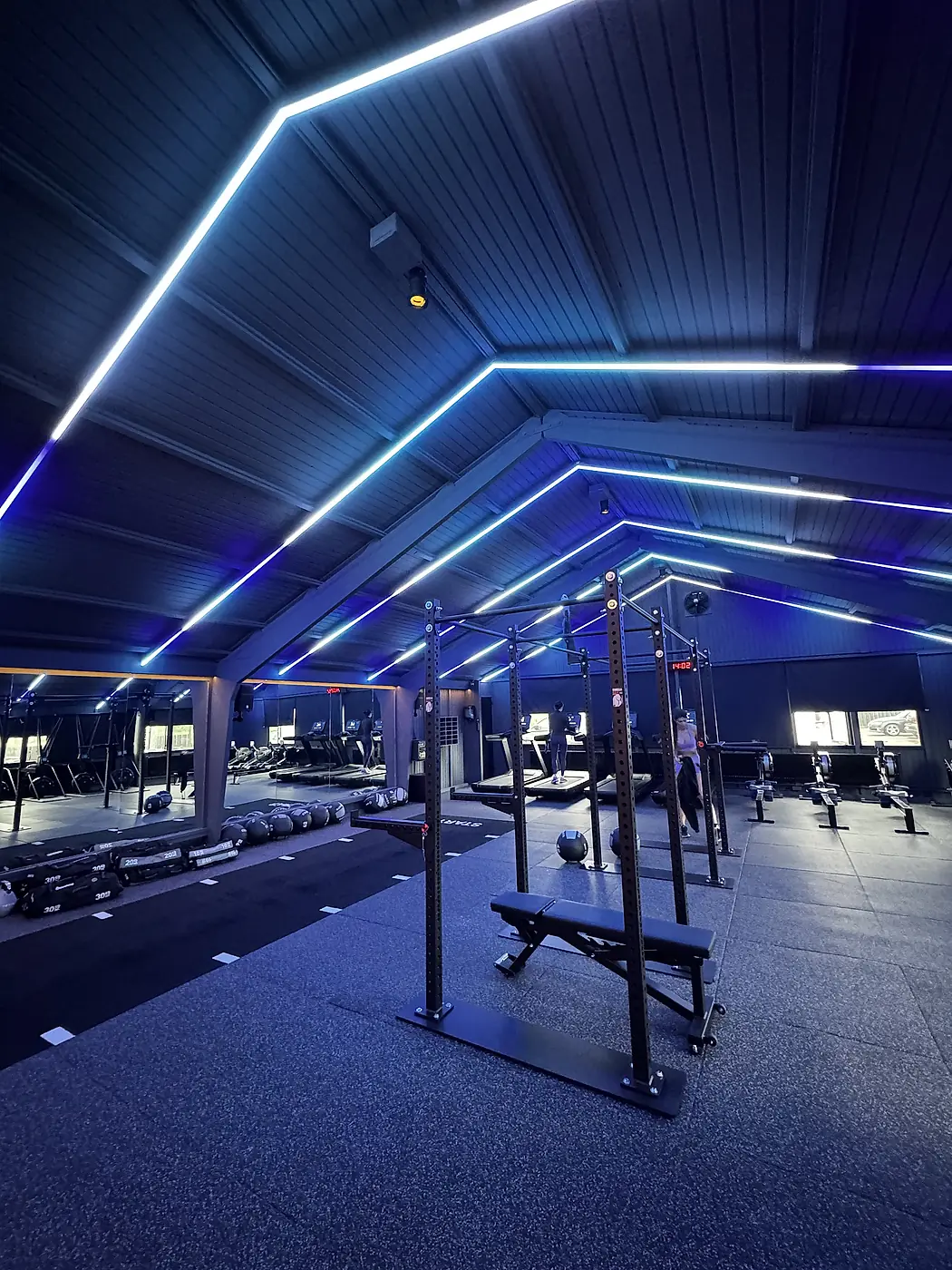 24UurFit Wilnes na oplevering: trainingsvloer met squat-racks, spiegelwand en blauwe LED-lijnen langs het houten puntdak